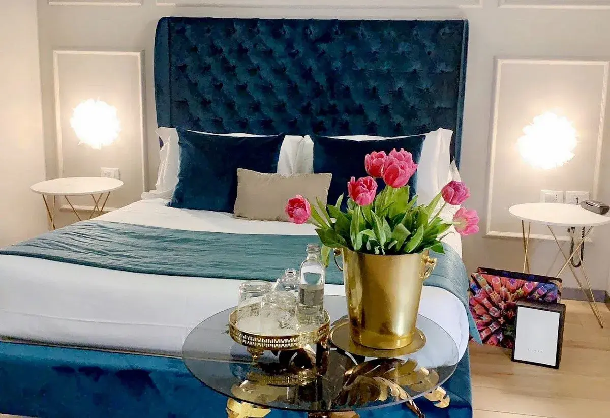 Chambre d'hôte en Lombardie : lit luxueux avec tête de lit capitonnée bleue, tulipes roses et tables de chevet élégantes.
