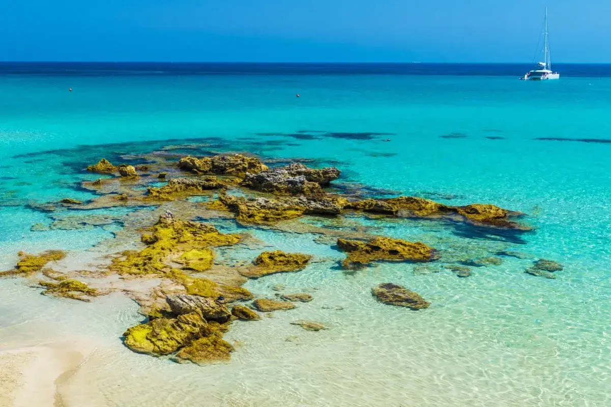 Eaux turquoise et rochers dorés sur une plage du sud de l'Italie. Un voilier blanc navigue au loin.