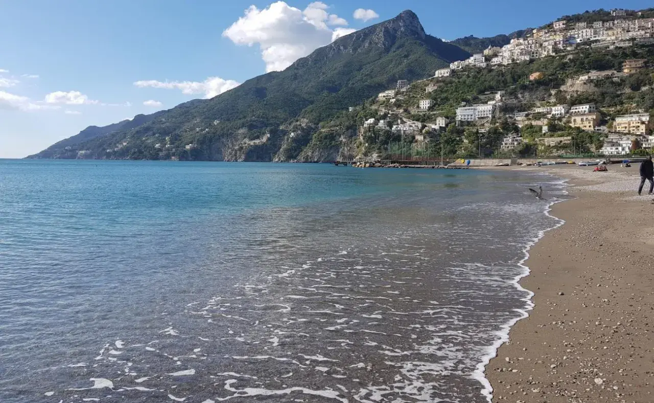 La plage de Vietri sul Mare s'étend sous un ciel bleu, avec des maisons blanches perchées sur la colline verdoyante. L'eau turquoise caresse le sable.