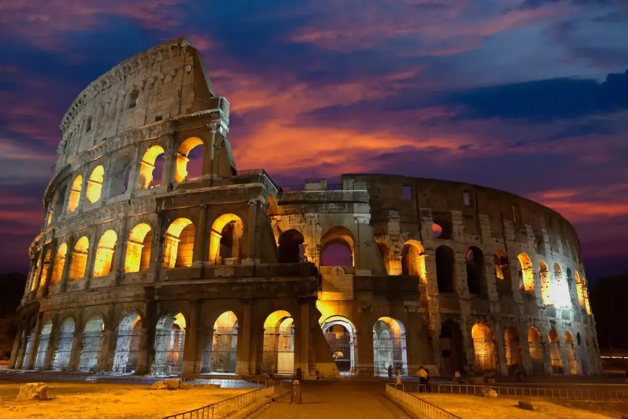 Le Colisée de nuit, illuminé par des lumières chaudes, se dresse sous un ciel aux couleurs flamboyantes.