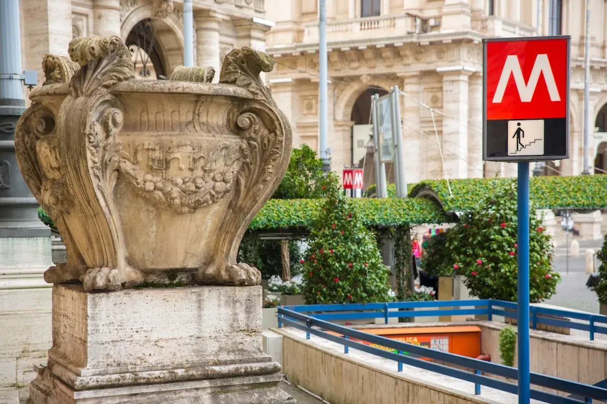 Un signe du **metro vatican** indique l'entrée d'une station, à côté d'une urne antique et de verdure luxuriante.