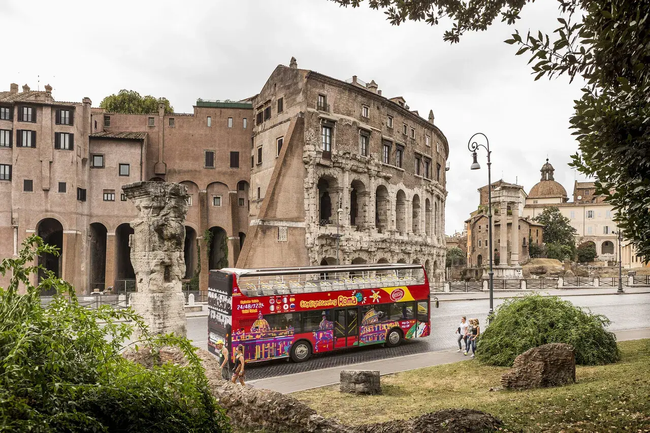 Un bus touristique rouge vif, "Citysightseeing Roma", passe devant des ruines antiques et des bâtiments romains.