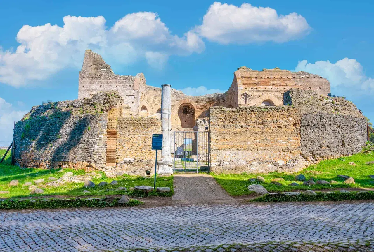 Ruines antiques sur l'Appia Antica à Rome, sous un ciel bleu parsemé de nuages blancs.