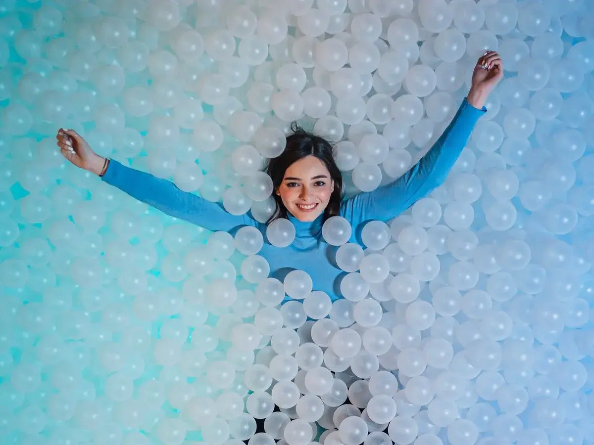 Une jeune femme souriante se détend dans une piscine de balles blanches, un clin d'œil ludique à l'ikono Rome.