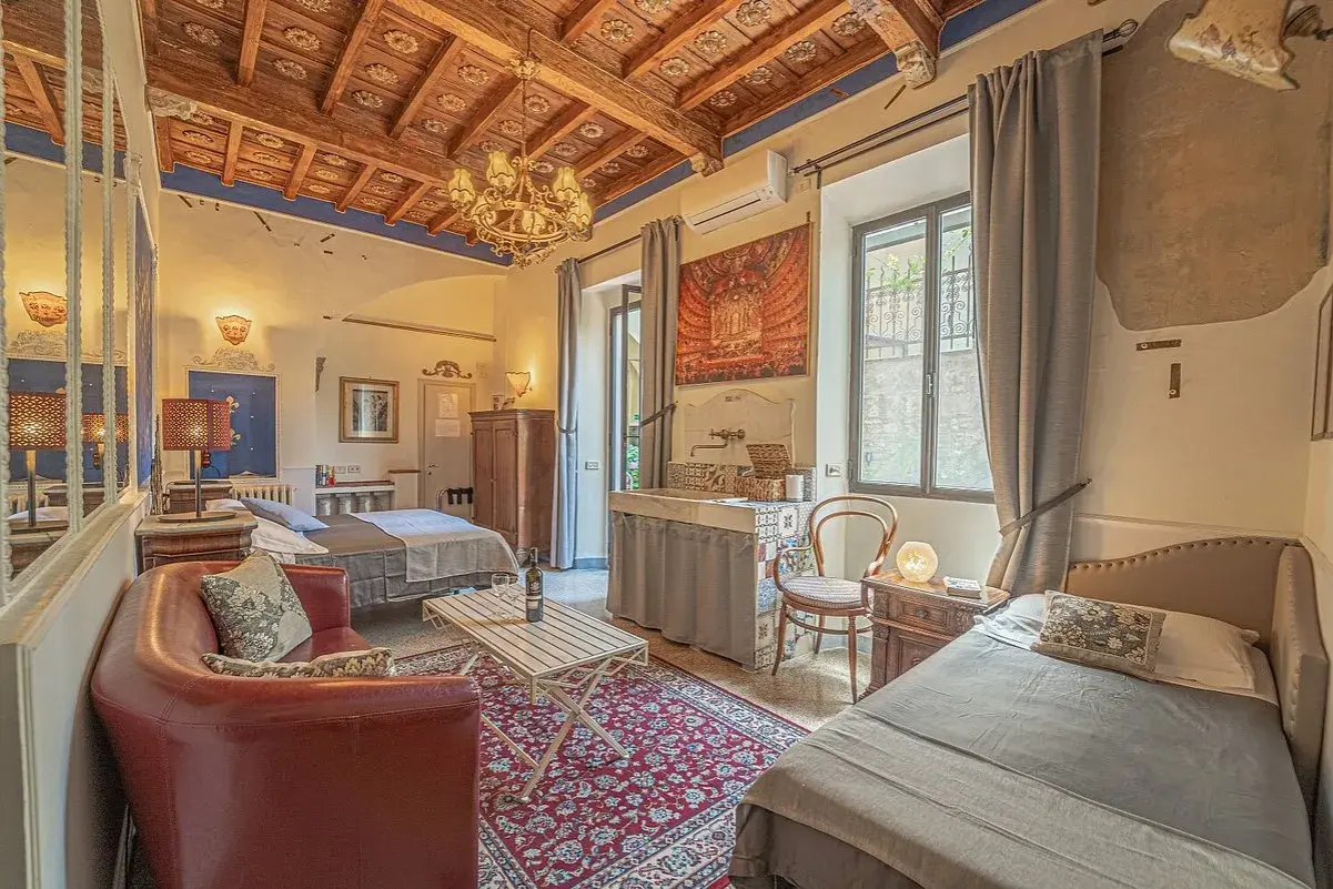 Chambre d'hôte toscane avec lits jumeaux, canapé rouge, tapis persan et plafond en bois sculpté.