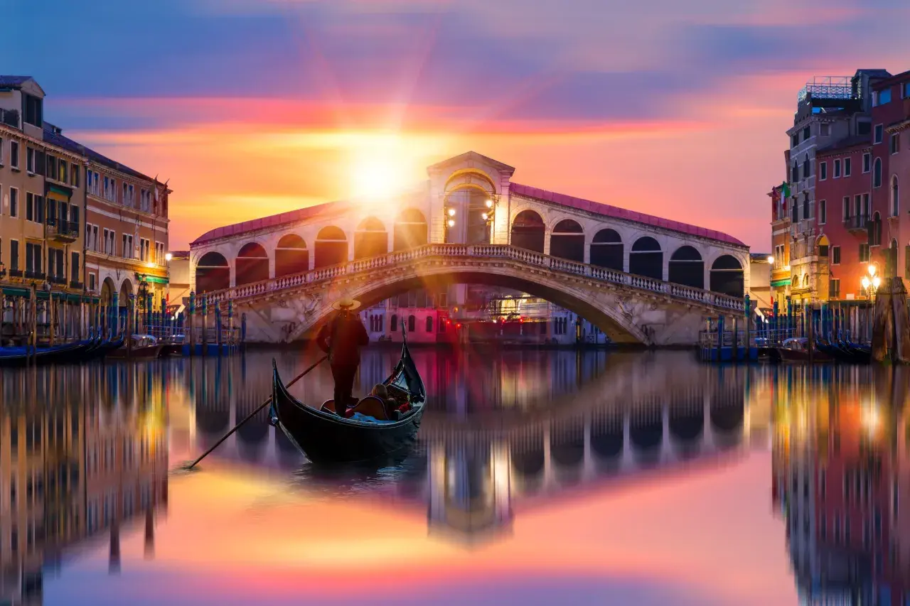 Gondole glissant sur le Grand Canal, sous un ciel aux couleurs vibrantes, avec le célèbre pont du Rialto en arrière-plan. Une scène qui évoque une célèbre peinture de Venise.