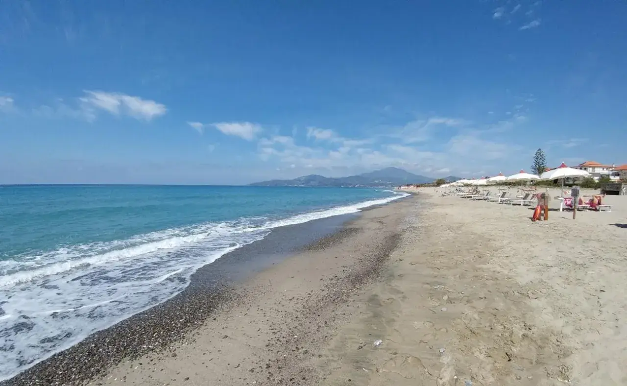 Plage de sable et de galets à Ascea Marina, avec des vagues bleues, des parasols blancs et des montagnes à l'horizon.