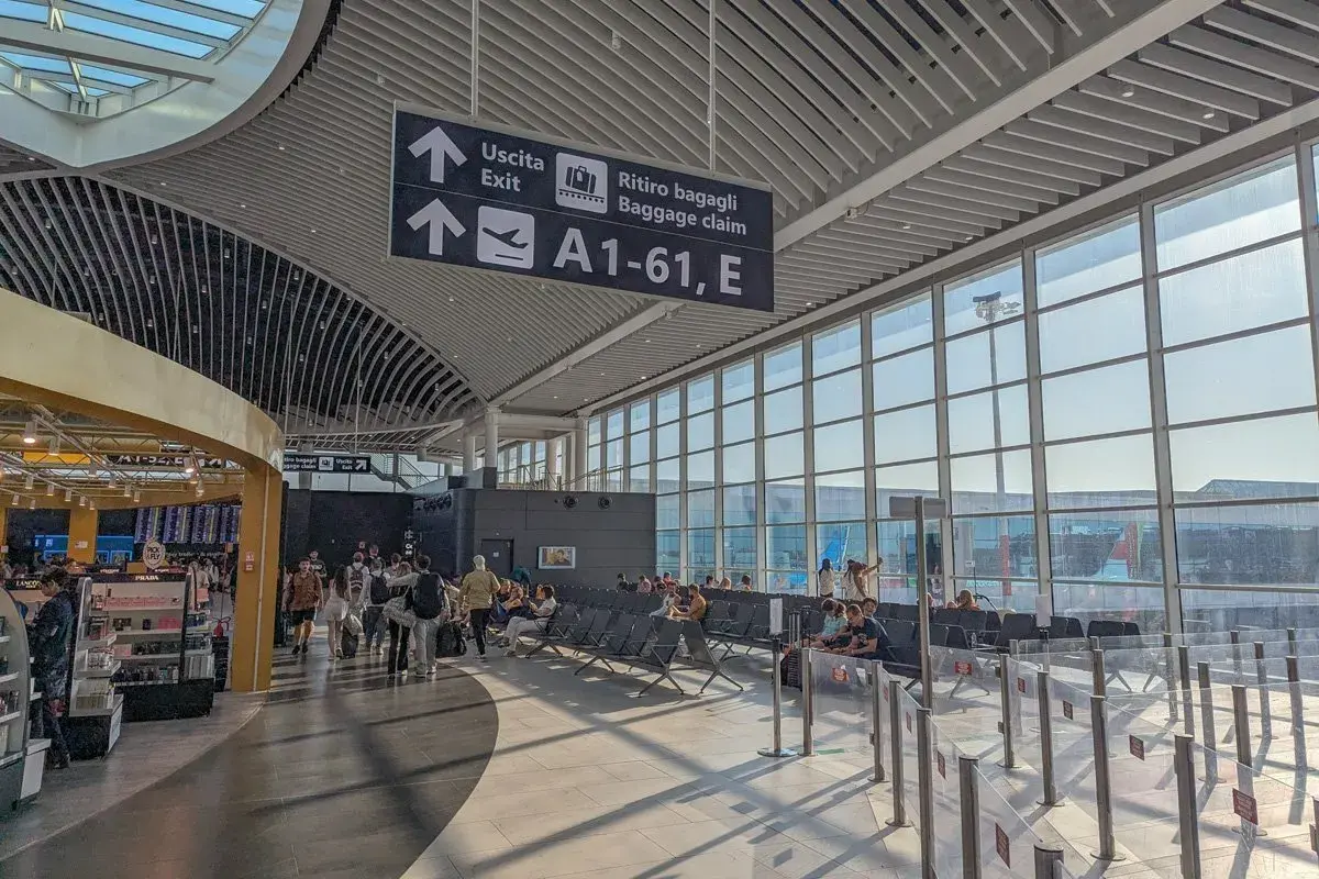 Intérieur lumineux d'un aéroport, avec des panneaux indiquant "Uscita" et "Ritiro bagagli". Des voyageurs attendent près des grandes baies vitrées, prêts pour leur vol au départ de l'aéroport Rome centre ville.