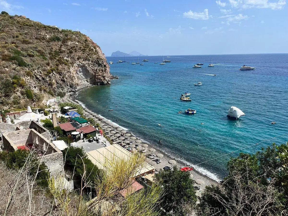 Plage animée à Lipari, avec des parasols, des petits restaurants et des bateaux naviguant sur la mer turquoise.