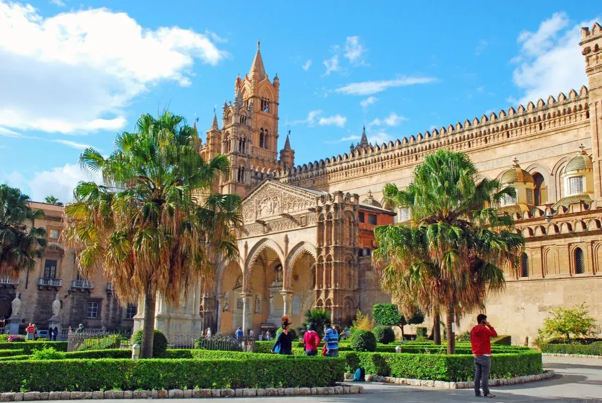 La cathédrale de Palerme, capitale Sicile, se dresse fièrement sous un ciel bleu, entourée de palmiers et de jardins verdoyants.