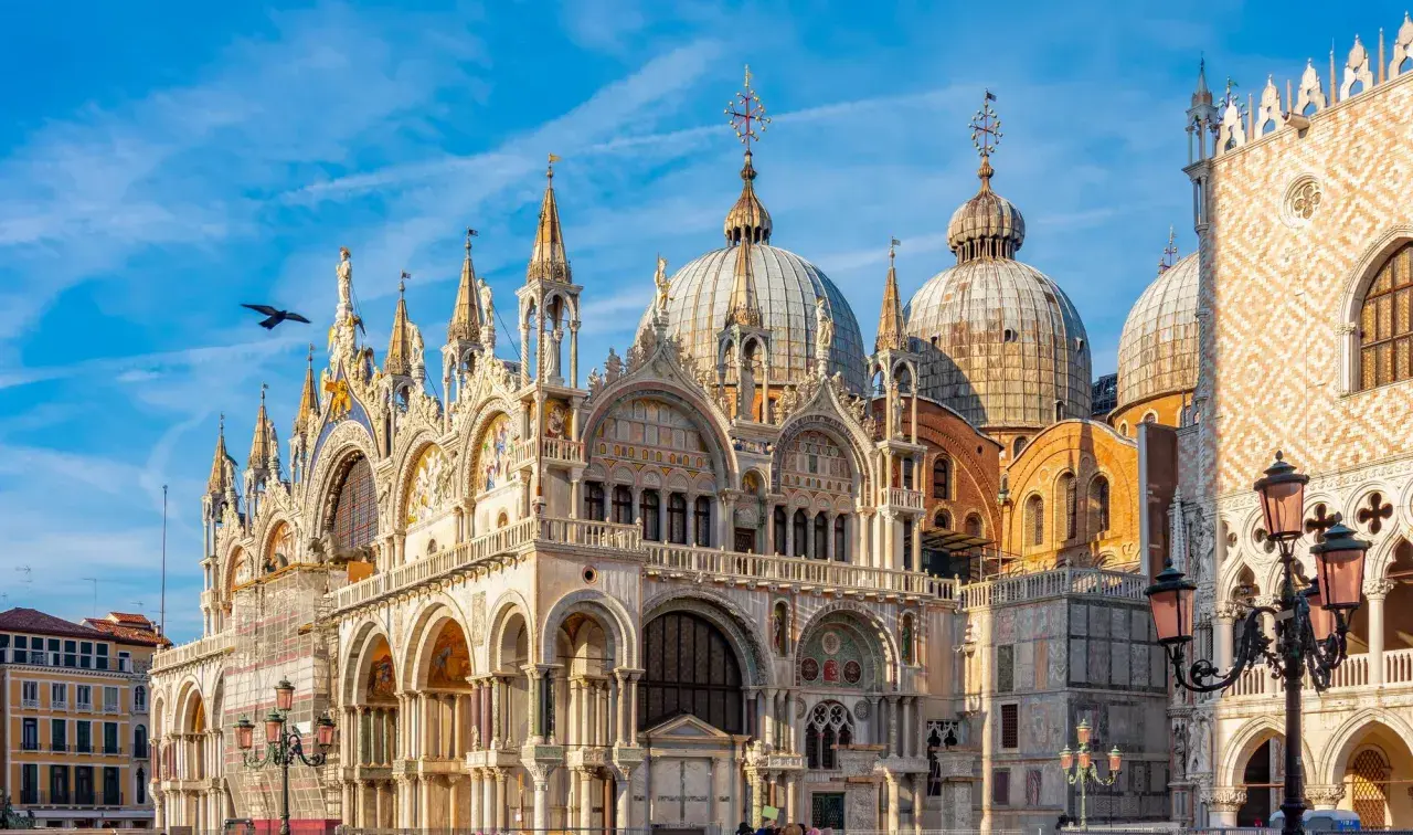 La basilique Saint-Marc à Venise, avec ses dômes majestueux et sa façade ornée, se dresse sous un ciel bleu éclatant. Un oiseau survole ce chef-d'œuvre architectural.