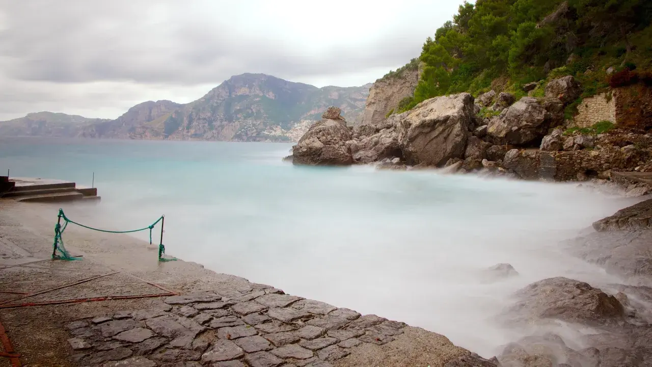 Vue brumeuse de la côte rocheuse de Praiano, Italie, avec des vagues douces et des montagnes en arrière-plan.