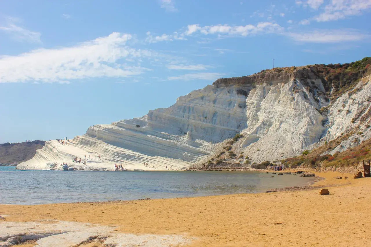 La Scala dei Turchi, une falaise blanche spectaculaire en Sicile, se dresse au-dessus d'une plage de sable doré et d'une mer bleue. Des visiteurs explorent ses terrasses naturelles.