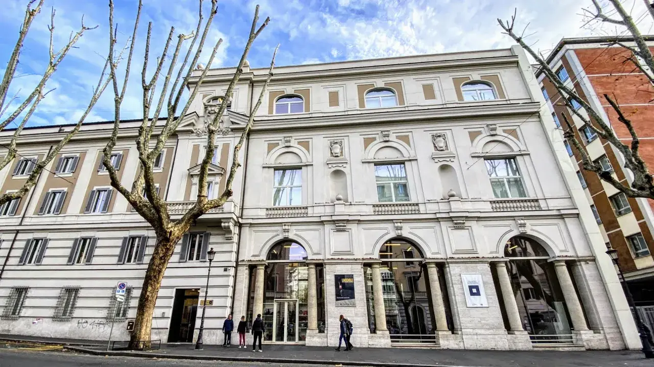 Le **Palazzo Merulana**, un édifice élégant, se dresse sous un ciel bleu nuageux, orné d'arbres dénudés aux branches graphiques.