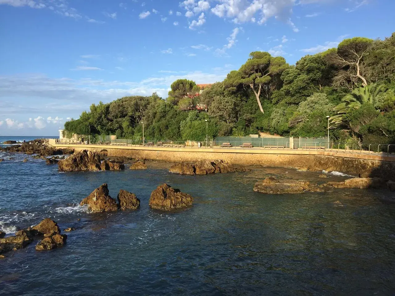 Promenade en bord de mer à Castiglioncello, Italie, avec des rochers dans l'eau et une colline verdoyante.