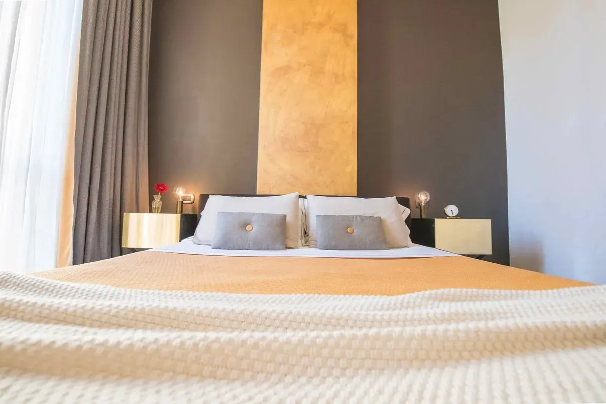 Lit et tables de chevet dorés dans une chambre d'hôte Italie du Nord. Rideaux gris et blancs, mur anthracite et ocre.