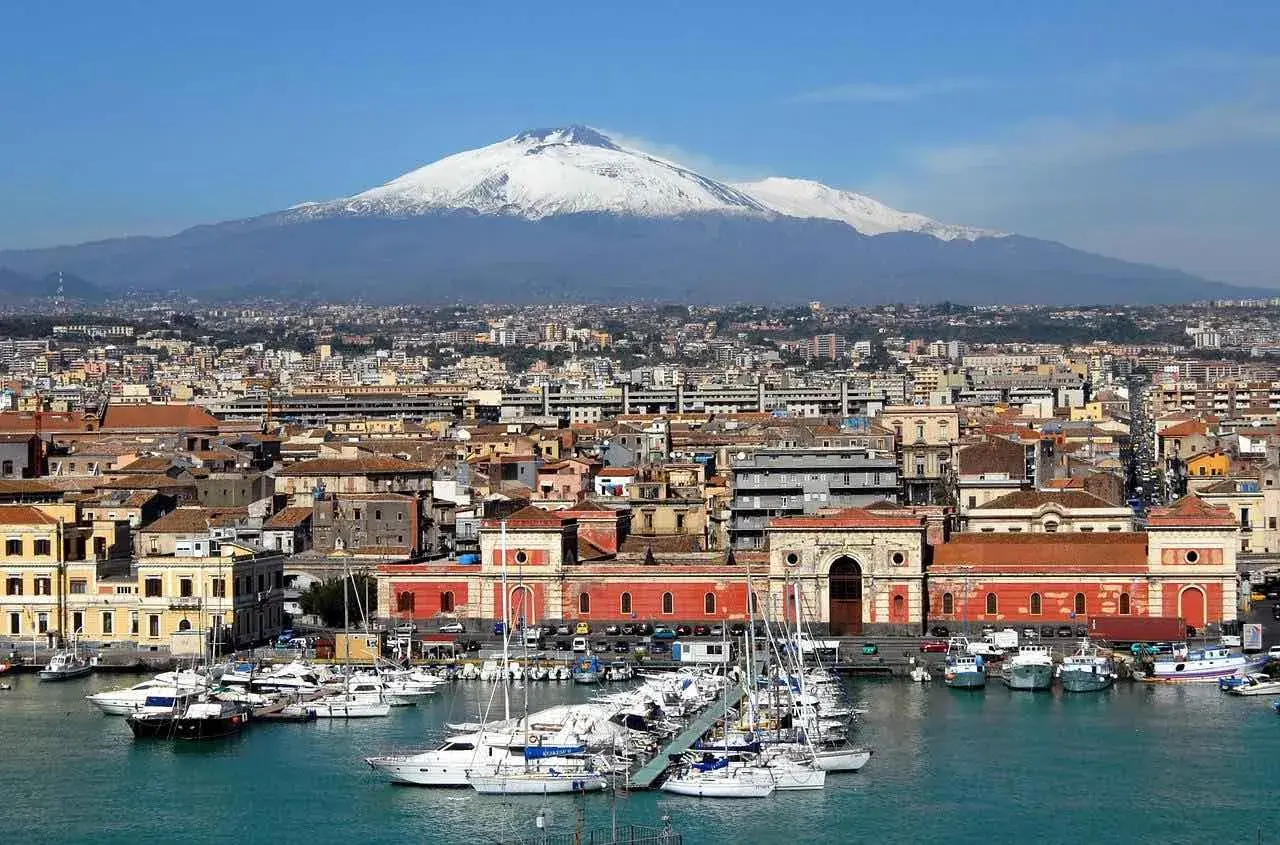 Vue panoramique de la ville de Catania, Sicile, avec le mont Etna enneigé en arrière-plan et un port rempli de voiliers.