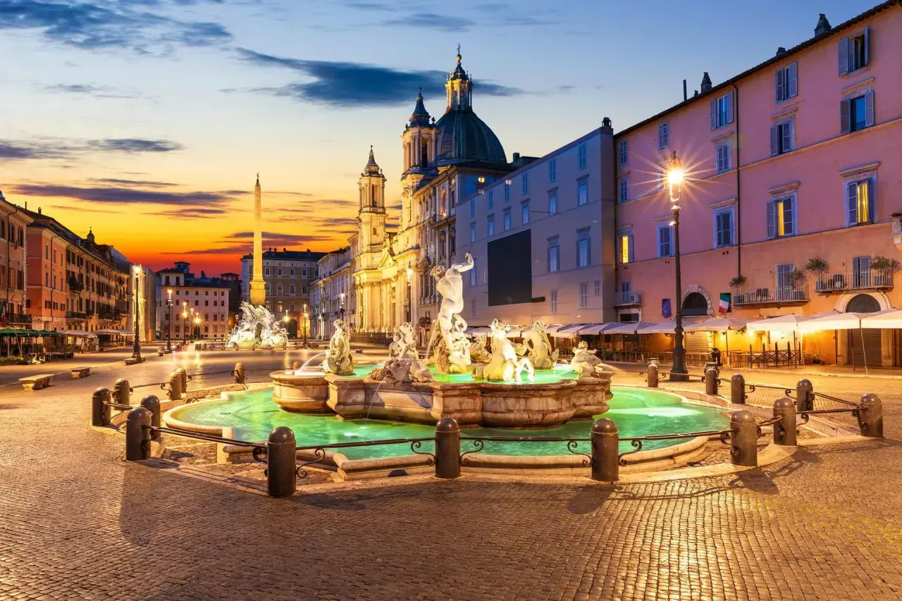 Fontaine de la piazza Navona illuminée au crépuscule, avec l'obélisque et l'église en arrière-plan.