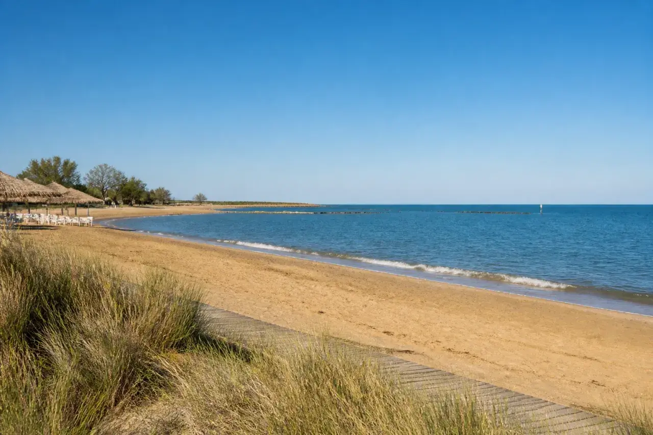 Plage de sable doré à Caorle, avec des parasols de chaume, des chaises blanches et la mer bleue sous un ciel clair.