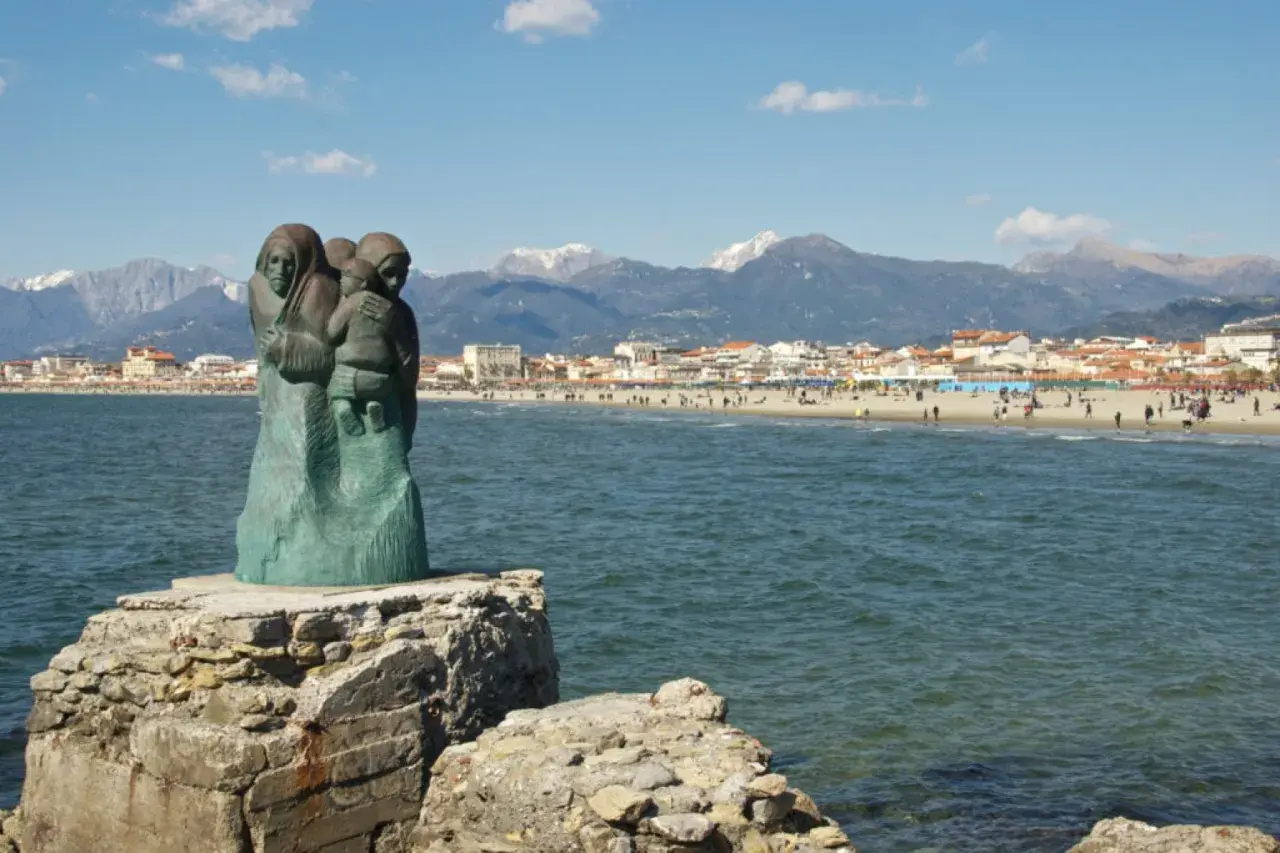 Sculpture de famille sur un rocher, face à la mer et à la ville de via Reggio, avec des montagnes enneigées en arrière-plan.
