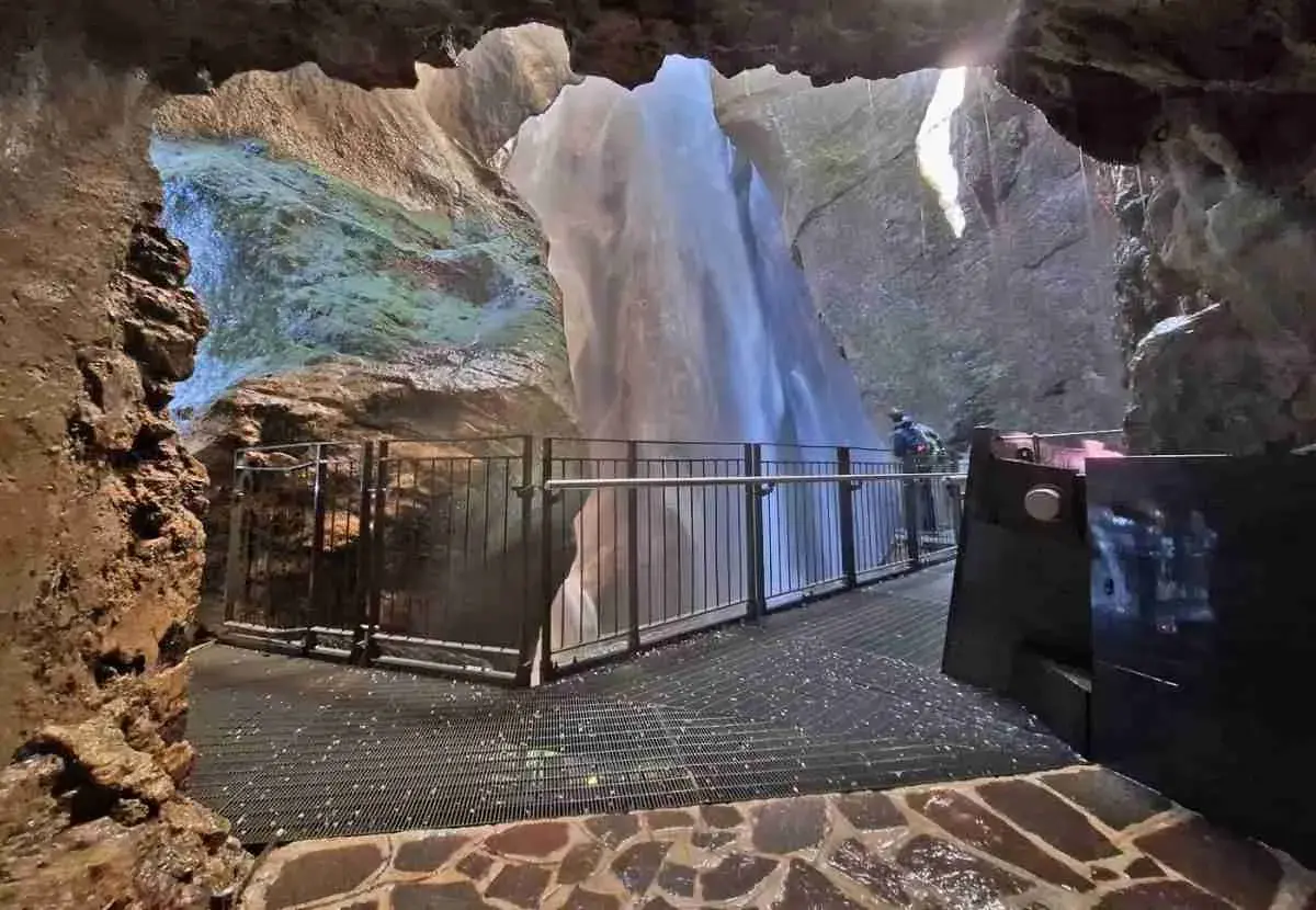 Vue depuis une grotte sur la cascade du Varone, avec une passerelle et un visiteur admirant la chute d'eau.
