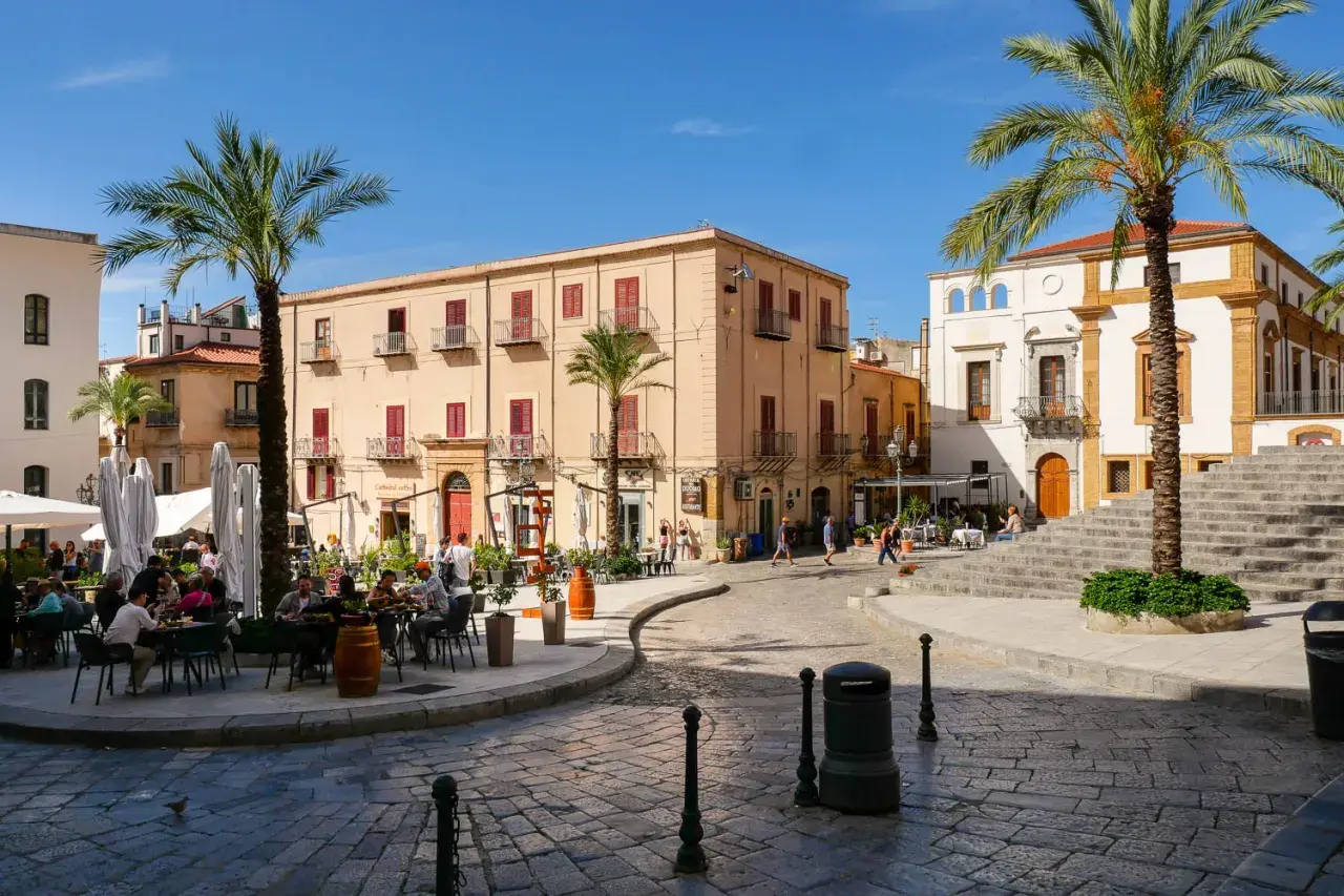 Place animée de Cefalù avec des terrasses de café, des palmiers et des bâtiments historiques sous un ciel bleu.