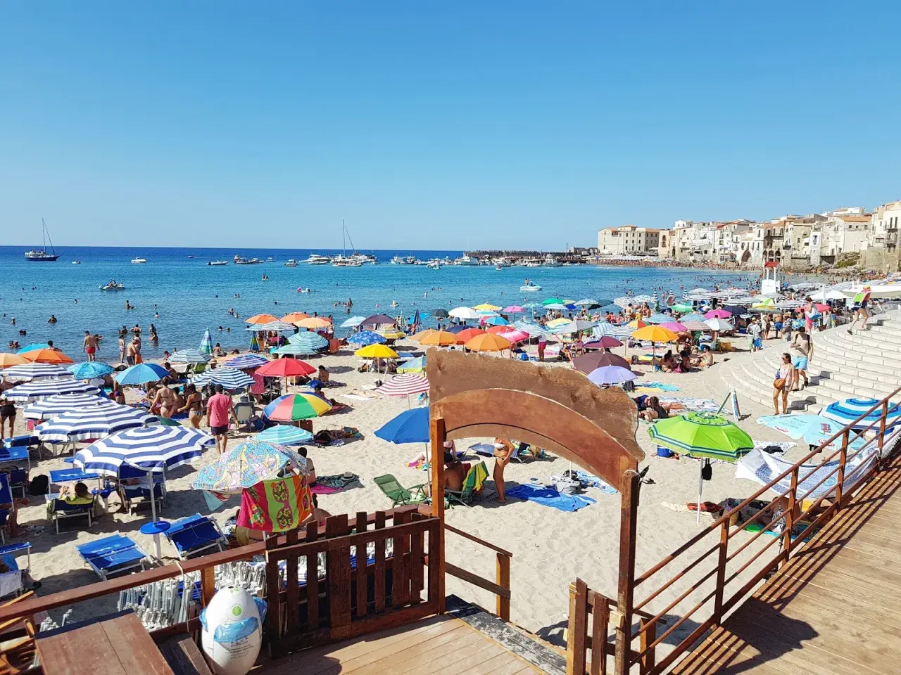 Plage animée à Cefalù en Sicile, avec des parasols colorés, des baigneurs dans la mer bleue et des bâtiments historiques en arrière-plan.