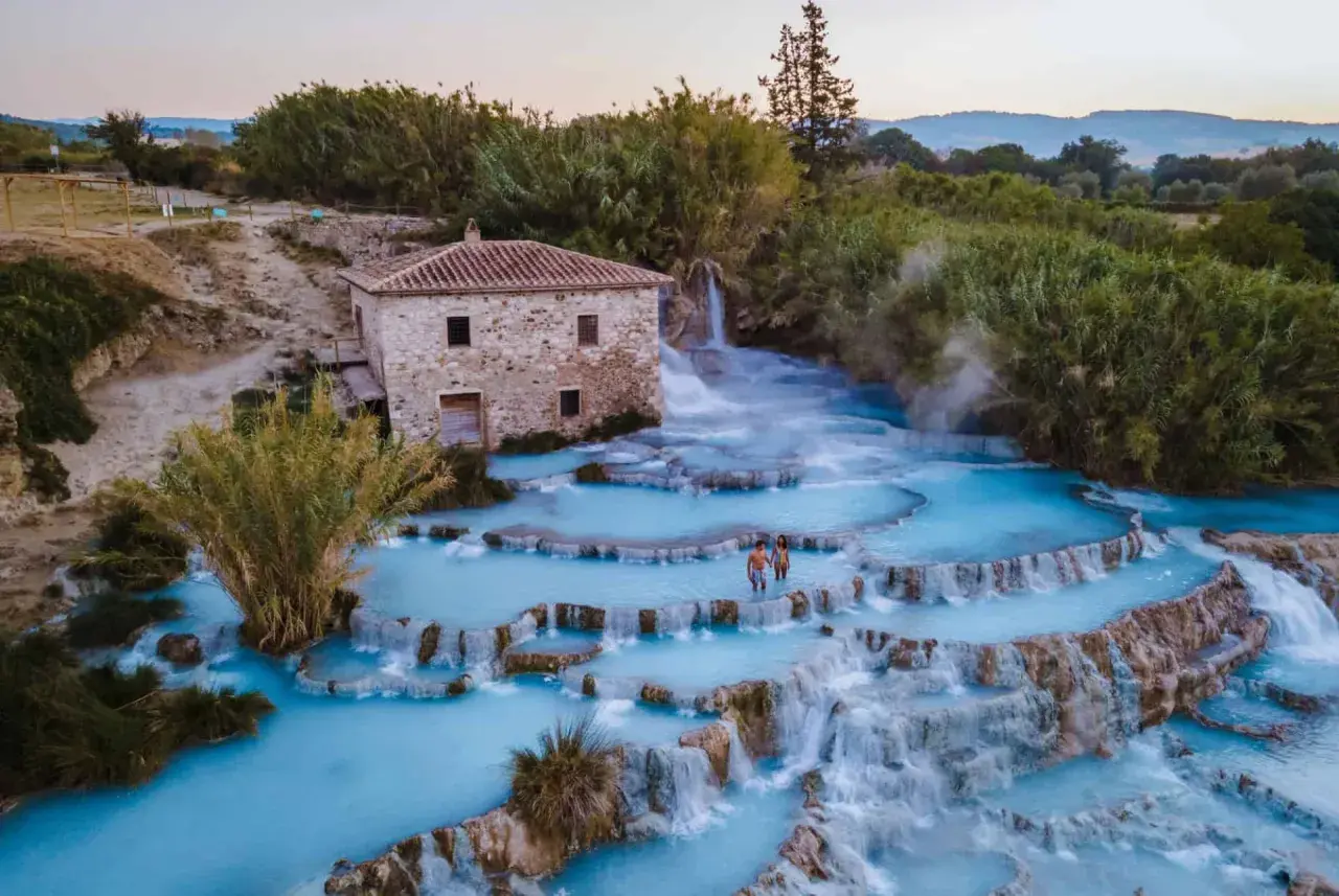 Casciana Terme : un couple se promène dans les cascades d'eau thermale bleue, près d'une vieille bâtisse en pierre.