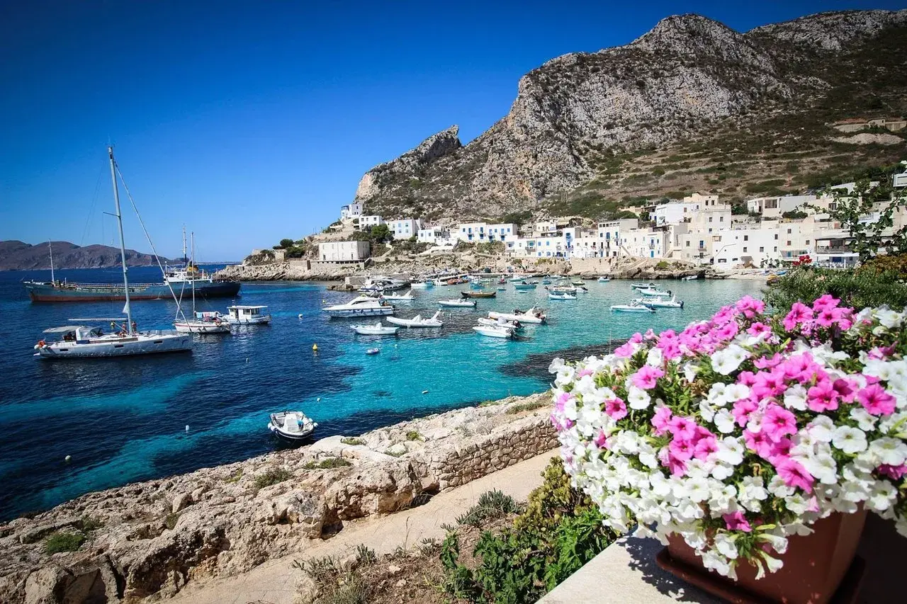 Vue d'un port pittoresque en Italie, avec des bateaux dans des eaux turquoise et des maisons blanches accrochées à flanc de colline. Un bouquet de fleurs roses et blanches encadre la scène, évoquant la beauté de Marsala, Italie.