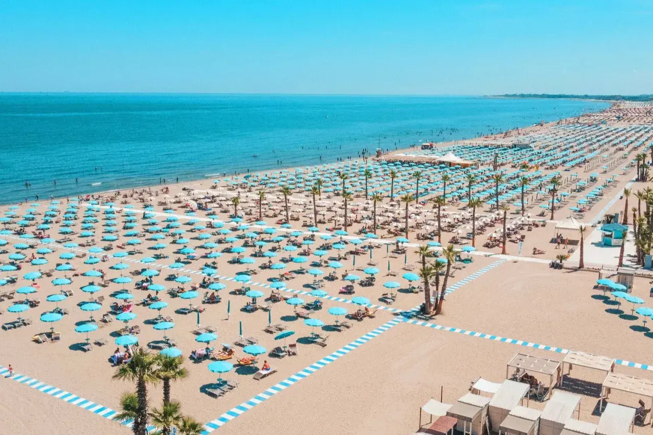 Plage de Chioggia, Italie, avec des rangées infinies de parasols bleus et blancs, des chaises longues et des palmiers sous un ciel bleu éclatant.