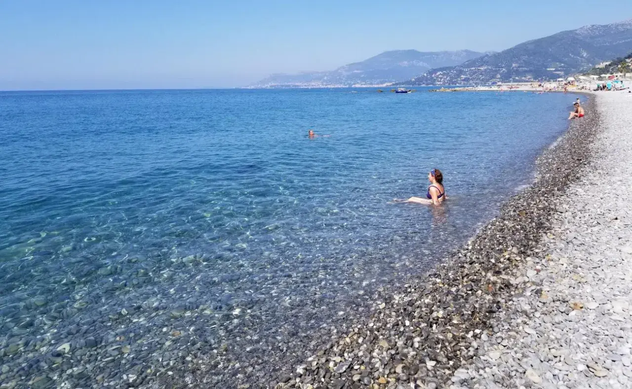 Une femme se baigne dans les eaux claires de la plage de galets de Ventimiglia, avec des montagnes en arrière-plan.
