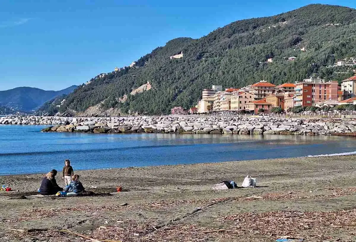 Vue de la plage de Chiavari avec des familles profitant du soleil, des bâtiments colorés et une colline verdoyante en arrière-plan.