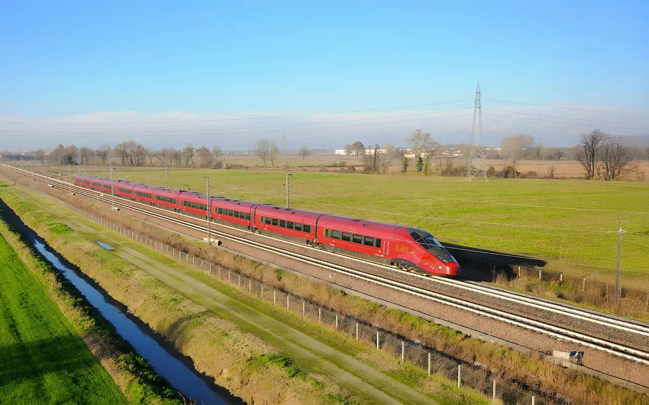Le train rouge Italo file sur les voies, reliant Roma à Naples à travers la campagne verdoyante sous un ciel bleu.