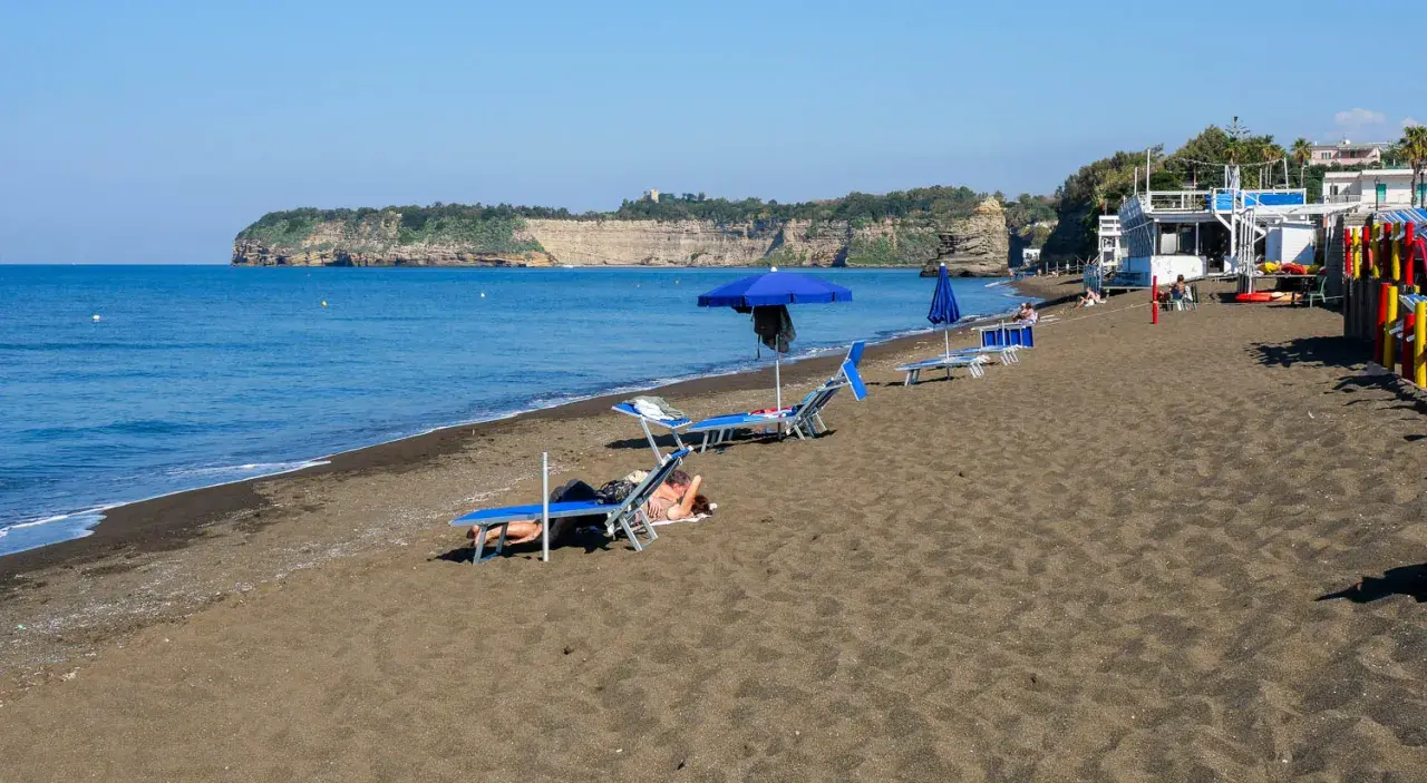 Plage de sable noir avec des chaises longues et des parasols bleus, face à une falaise rocheuse. Un doux air de Procida ou Capri.