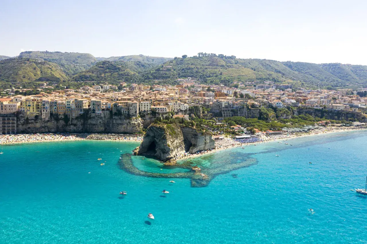 Vue aérienne de Tropea, avec ses maisons colorées perchées sur la falaise, une plage bondée et la mer d'un bleu turquoise éclatant.