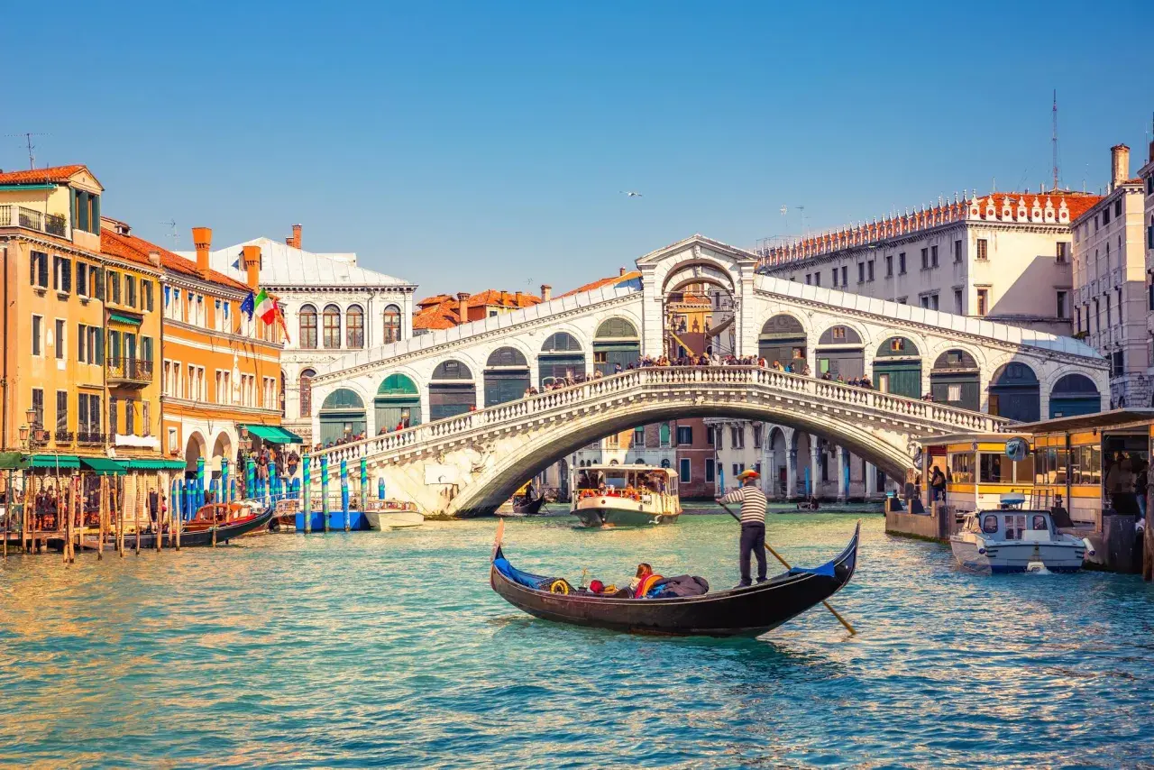 Gondole naviguant sur le Grand Canal, sous le célèbre pont du Rialto à Venise. L'eau turquoise reflète les façades colorées des bâtiments.