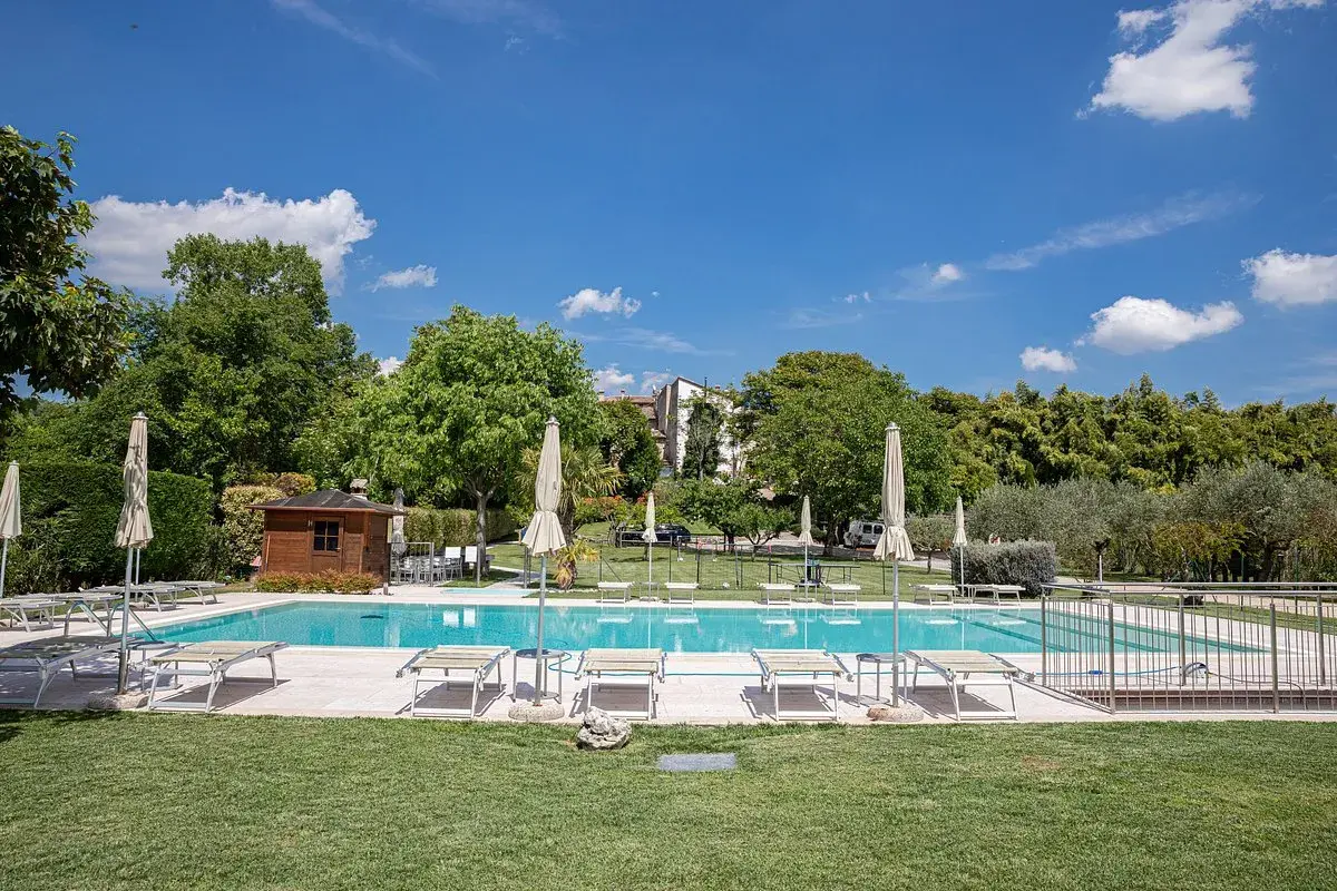 Piscine d'agritourisme au Lac de Garde, entourée de verdure, avec chaises longues et parasols sous un ciel bleu.