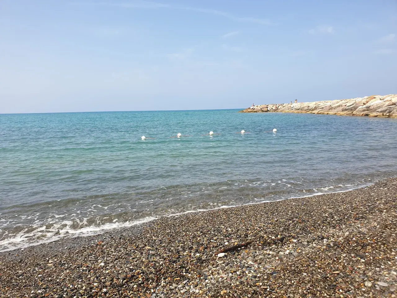 Plage de galets à Cecina Mare, avec des bouées blanches marquant la zone de baignade et une jetée rocheuse au loin.