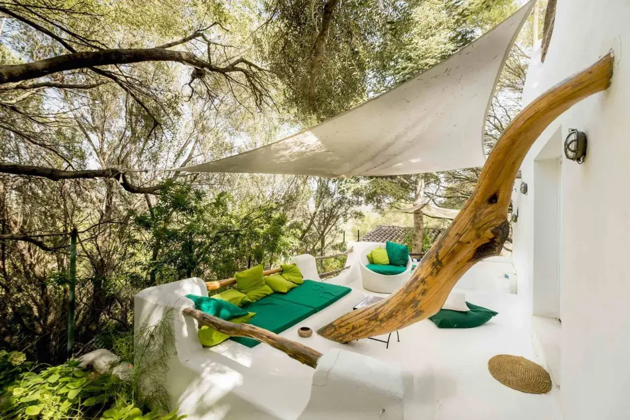 Terrasse du Hotel Su Gologone avec mobilier blanc et coussins verts, sous une voile d'ombrage et entourée de verdure.