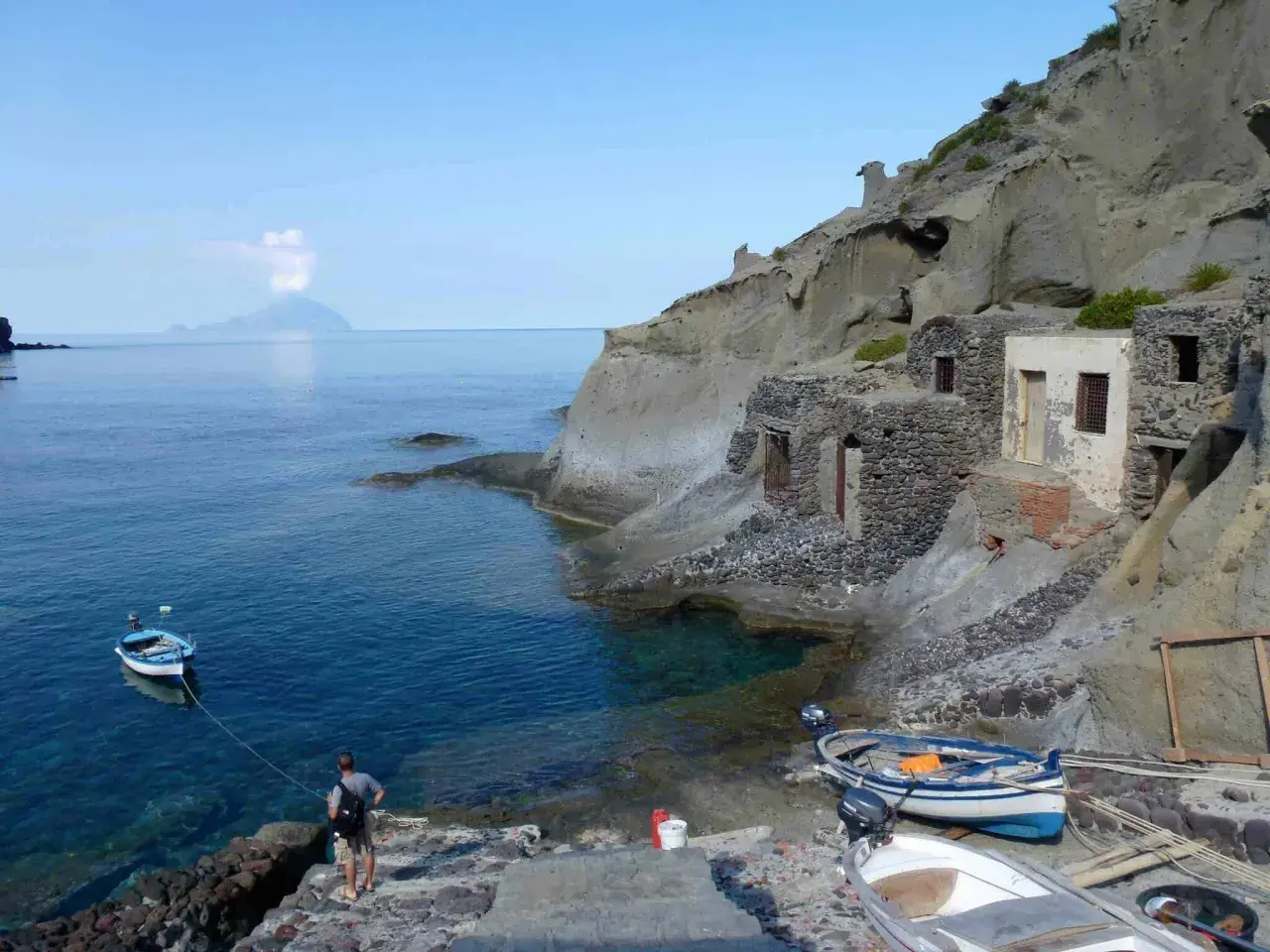 Sur l'île Salina, un homme amarre un bateau près de vieilles maisons de pêcheurs en pierre, avec une île volcanique au loin.