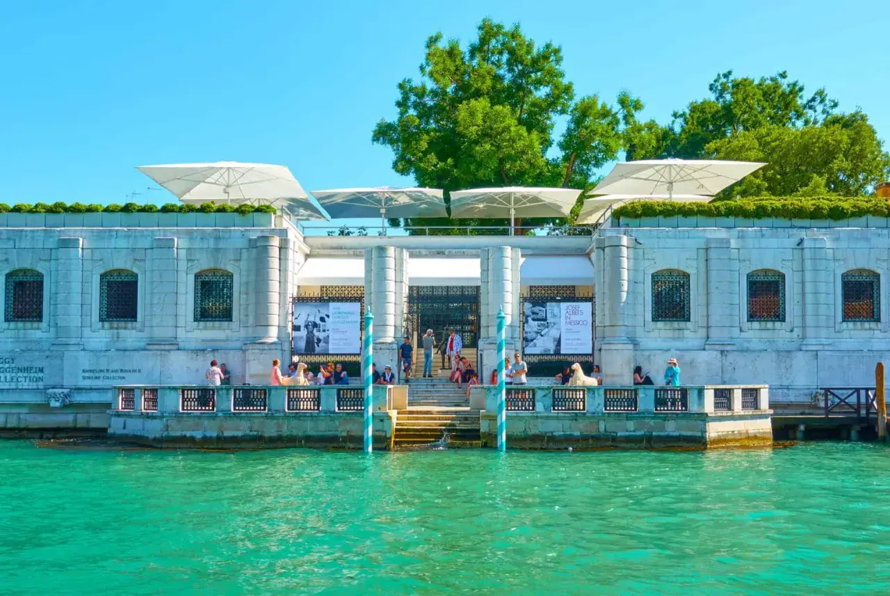 Le **Guggenheim Venise** se reflète dans l'eau turquoise. Des parasols blancs ombragent la terrasse où des visiteurs admirent l'architecture.