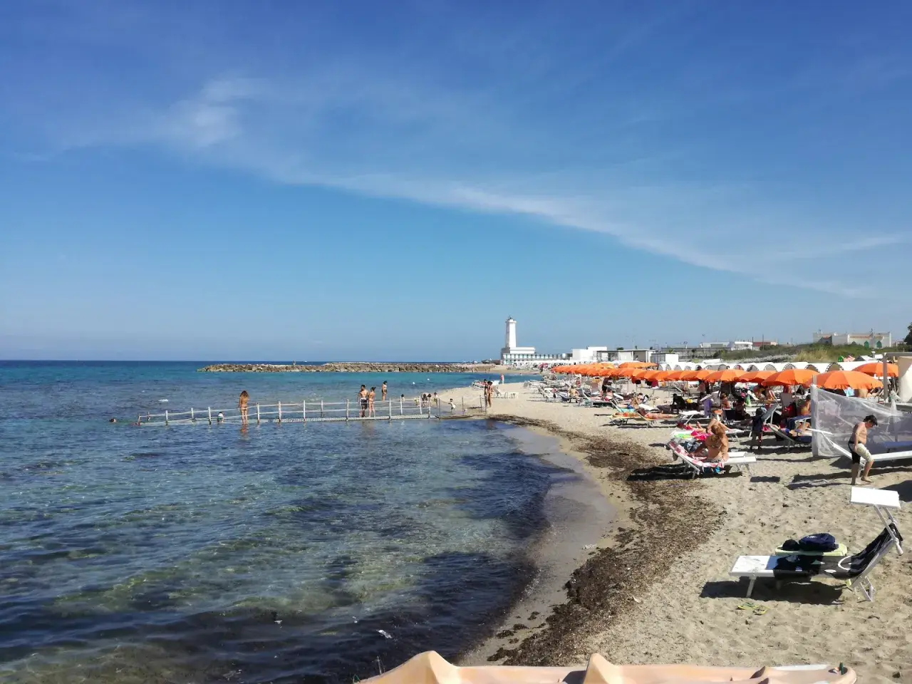 Plage animée à San Cataldo, Pouilles, avec des parasols orange, des baigneurs sur un ponton et un phare blanc en arrière-plan.