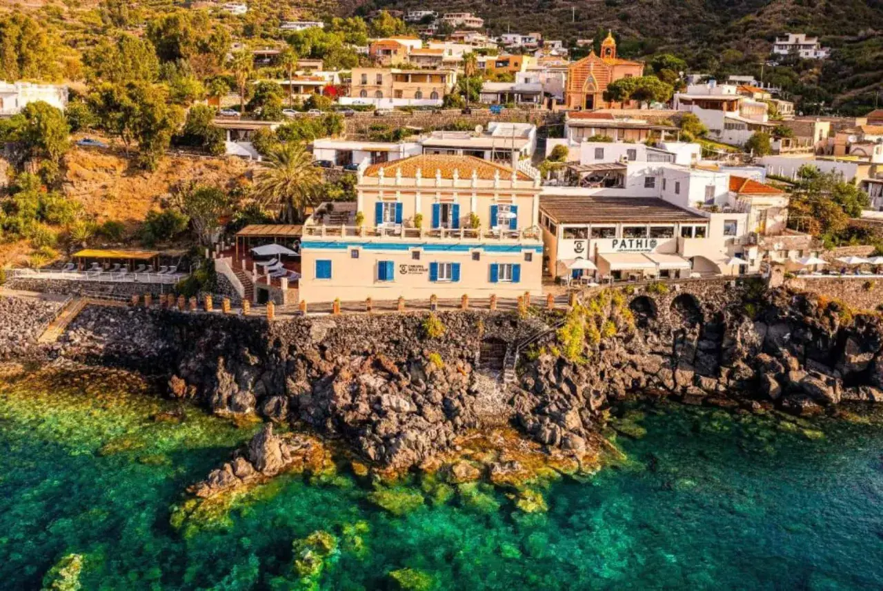 Vue d'un gîte en Sicile, perché sur une falaise rocheuse surplombant une mer turquoise. Bâtiments blancs et ocres, végétation luxuriante.