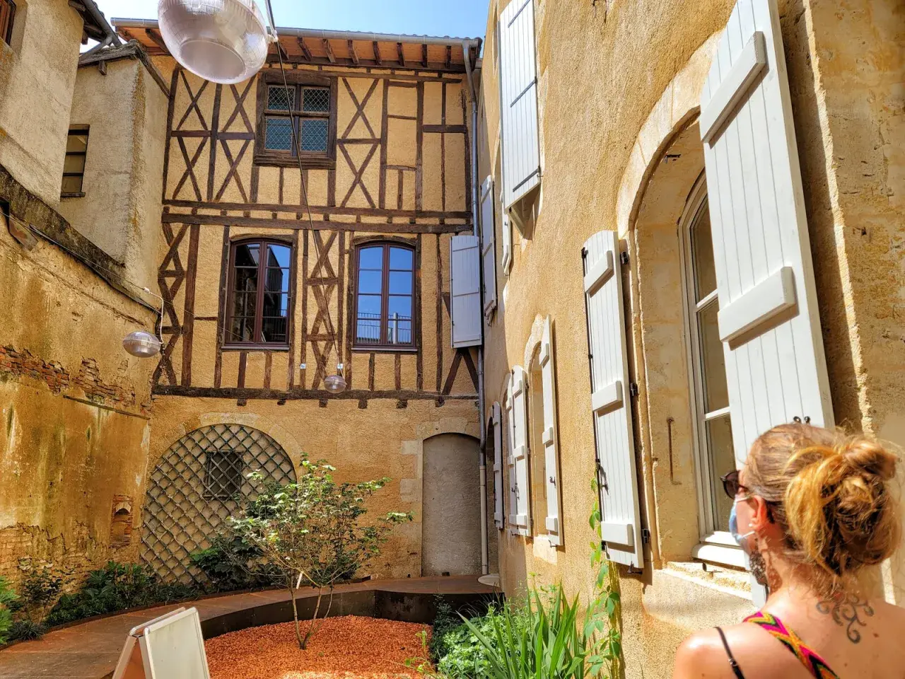 Cour intérieure avec colombages et volets blancs. Une femme regarde la scène, comme si elle découvrait un coin charmant de Fleurance, Italie.