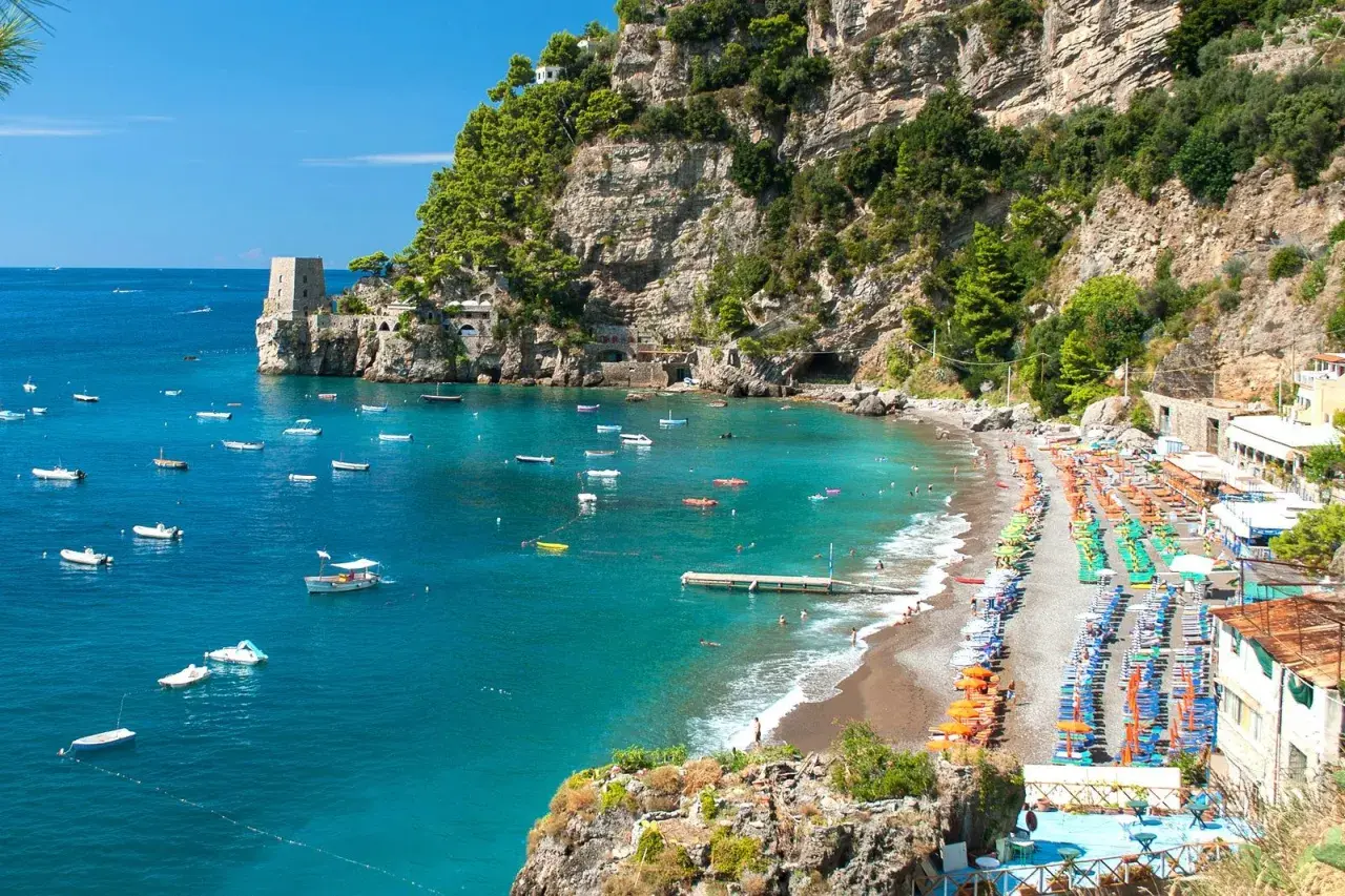 Vue panoramique d'une plage de galets animée à Almafi, Italie, avec des parasols colorés, des chaises longues et des eaux bleues claires parsemées de bateaux.