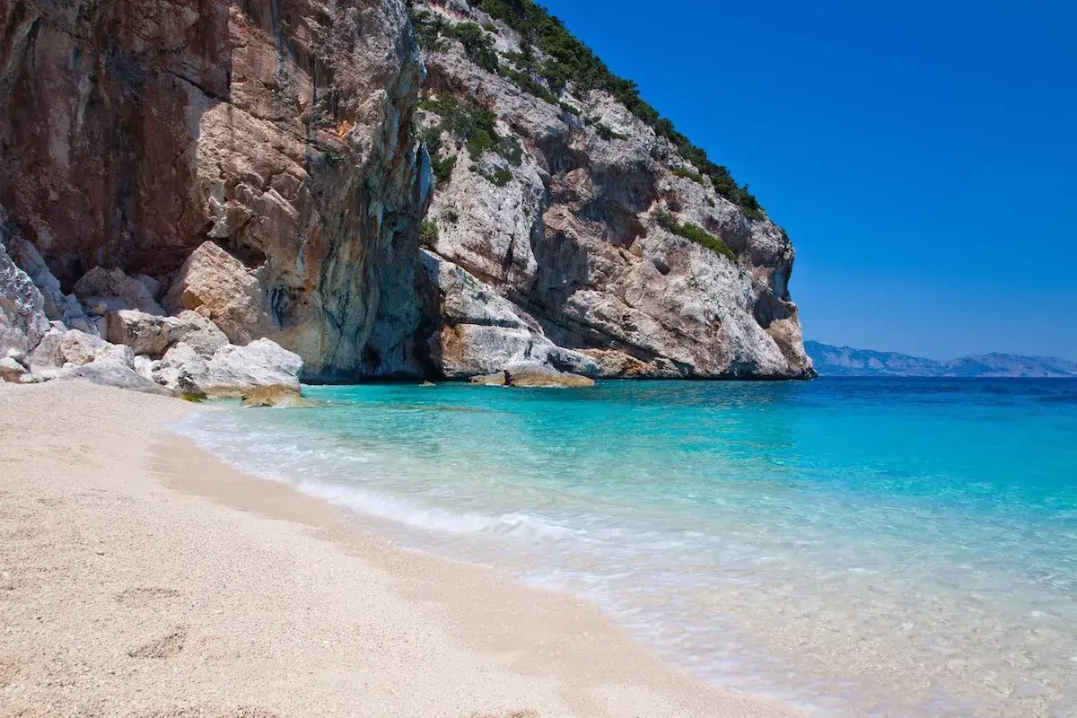 Plage de sable fin bordée par une eau turquoise cristalline, avec une falaise imposante en arrière-plan. Un coin de paradis sur la côte smeralda sardaigne.