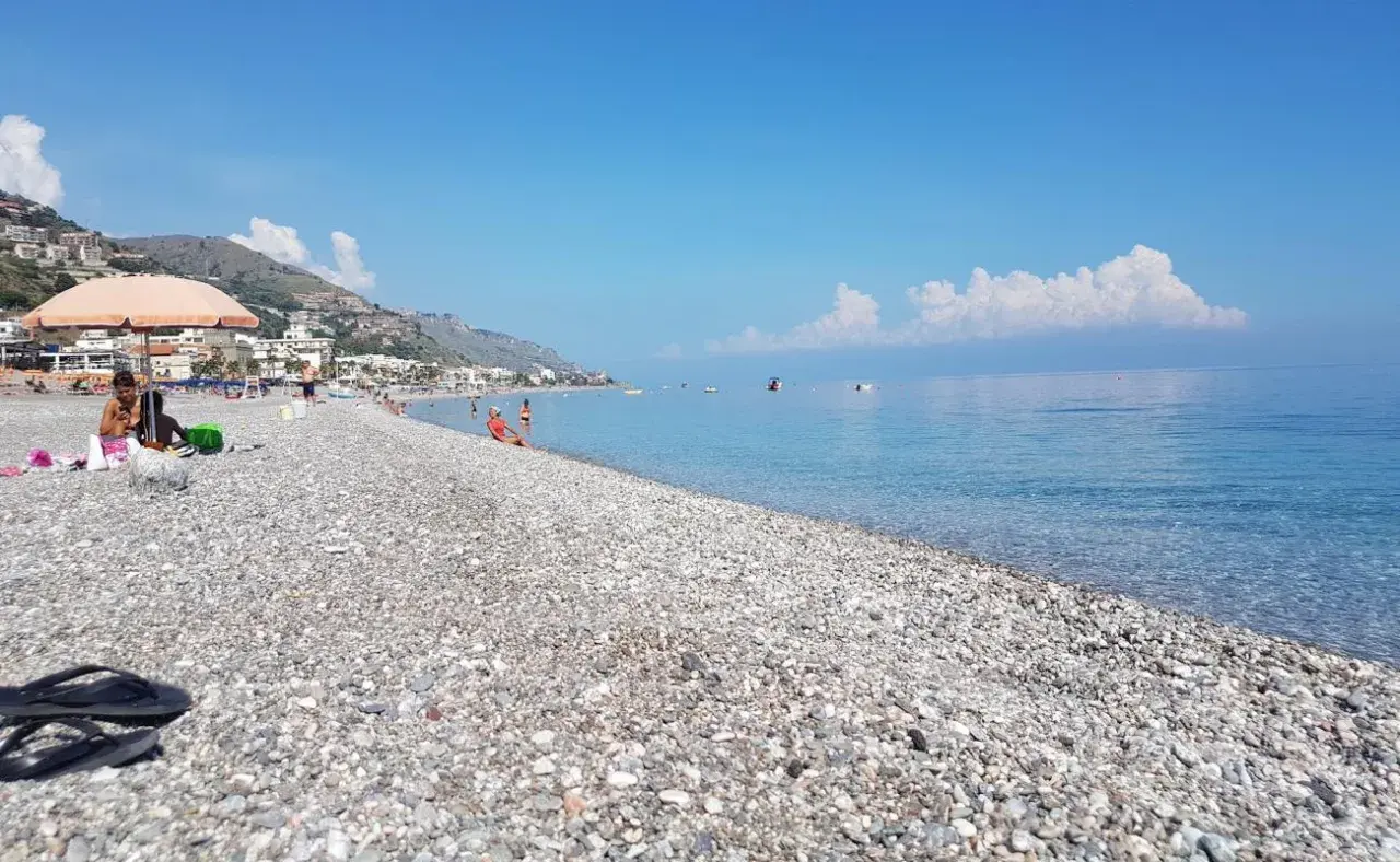 Plage de galets à Letojanni, Sicile. Des baigneurs profitent du soleil sous un parasol, tandis que la mer d'un bleu profond s'étend à l'horizon.