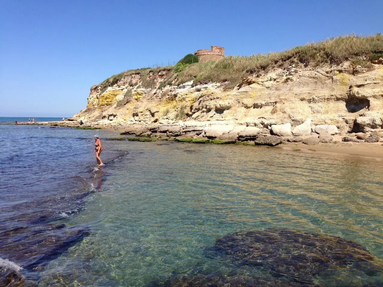 Une femme en bikini profite de la mer limpide près d'une falaise rocheuse à Anzio, avec une tour ancienne en arrière-plan.