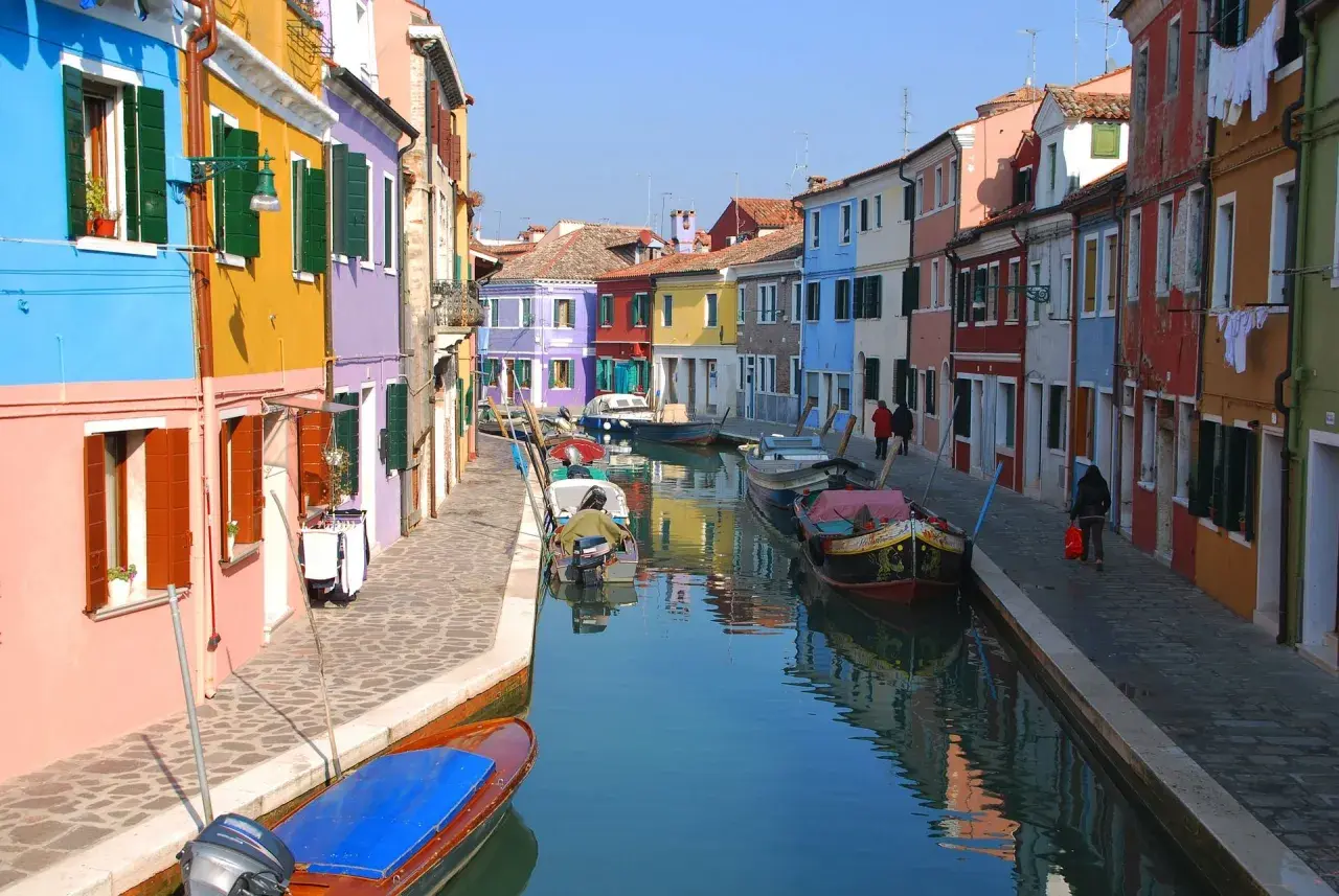Canal bordé de maisons colorées à Murano, Venise. Des bateaux sont amarrés le long de la voie navigable, reflétant les façades vibrantes.
