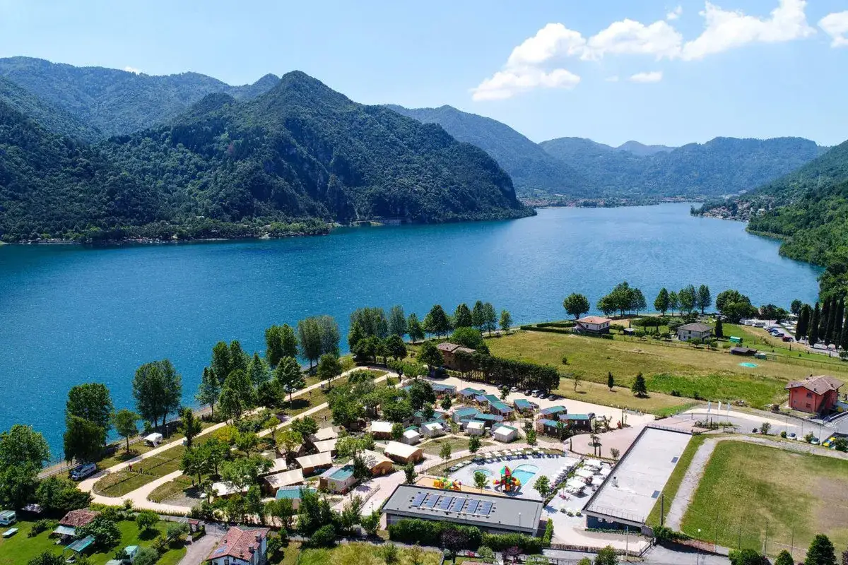 Vue aérienne du lac d'Idro, entouré de montagnes verdoyantes. Un camping avec piscine et bungalows est visible au premier plan.